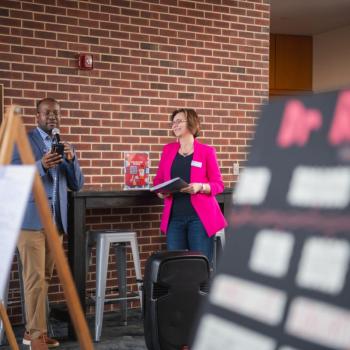 Two presenters speak near poster displays on easels in a brick-walled indoor atrium.