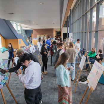 Crowded poster session with attendees viewing displays on easels in a sunlit atrium.