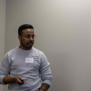 Person giving a presentation in a room, wearing a gray sweater with a name tag, holding a small device in one hand.